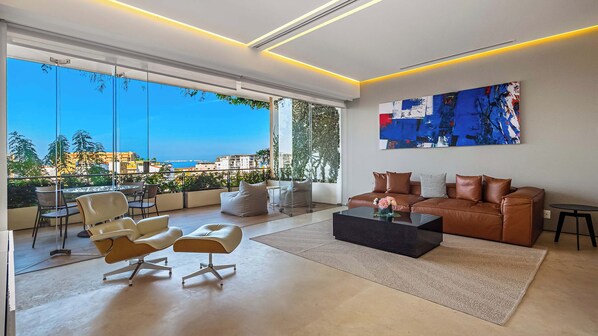 A modern living room with a large window, a brown leather sofa, a black coffee table, and a painting on the wall.