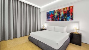A modern hotel room with a large bed, two bedside tables with lamps, a colorful abstract painting on the wall, and grey curtains.