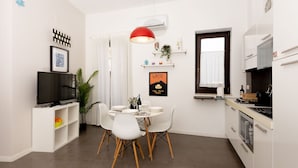 A compact kitchen with white cabinets, a dining area with a round table and chairs, a TV on a stand, and a red pendant light.