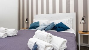 A neatly made bed with white and blue pillows, a purple quilt, and rolled white towels on a wooden nightstand.