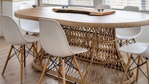 Une table de salle à manger ronde, entourée de chaises blanches dont le dossier est en osier, dans une pièce dotée de grandes fenêtres et d’un ventilateur de plafond.