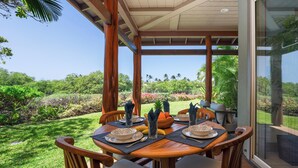 A dining area with a table set for four, surrounded by wooden chairs with cushioned seats. The table is placed under a wooden pergola with a glass door, offering a view of a lush garden and distant hills. - Mauna Lani Beach Club Pass included!  701 Fairways at Mauna Lani (Mauna Lani Resort)