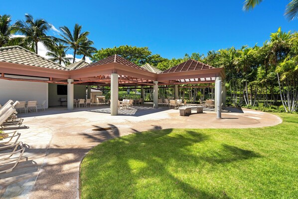 A covered outdoor patio with seating and a pool area, surrounded by palm trees and a clear sky.