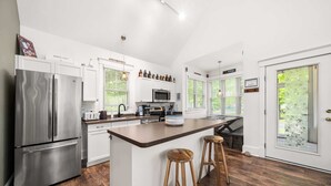 A modern kitchen with a large island, stainless steel appliances, and wooden stools. - The Trillium - 2 Pine Forest (Prince Edward)