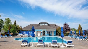 Una piscina con lettini e ombrelloni, un edificio con facciata blu e alberi sullo sfondo.