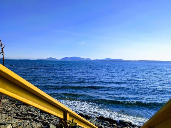 Ein Strand mit einem hölzernen Surfbrett, das an Felsen lehnt, ruhigem Meer und Bergen in der Ferne.