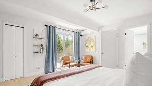 A bedroom with a large bed, two chairs, a small table, a window with curtains, and a door. - Bright & Modern | King Suite | Creekside Trail (Boulder)