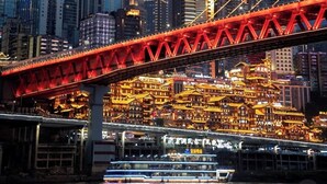 Stadsgezicht met een opvallende rode brug en verlichte gebouwen, waaronder een hoge wolkenkrabber met verticale lichten.