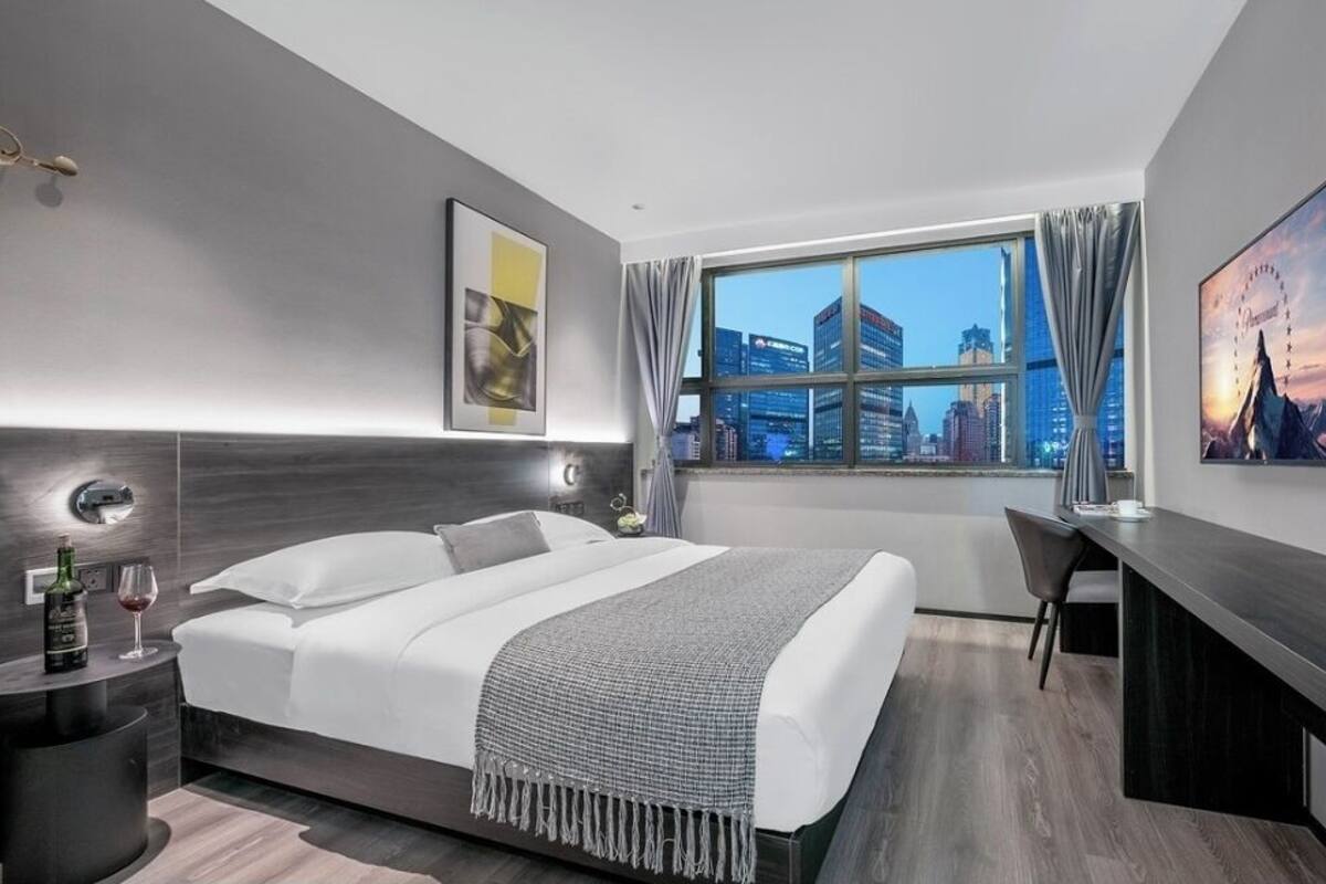 A modern hotel room with a large bed, a desk, and a view of the city skyline through a large window.