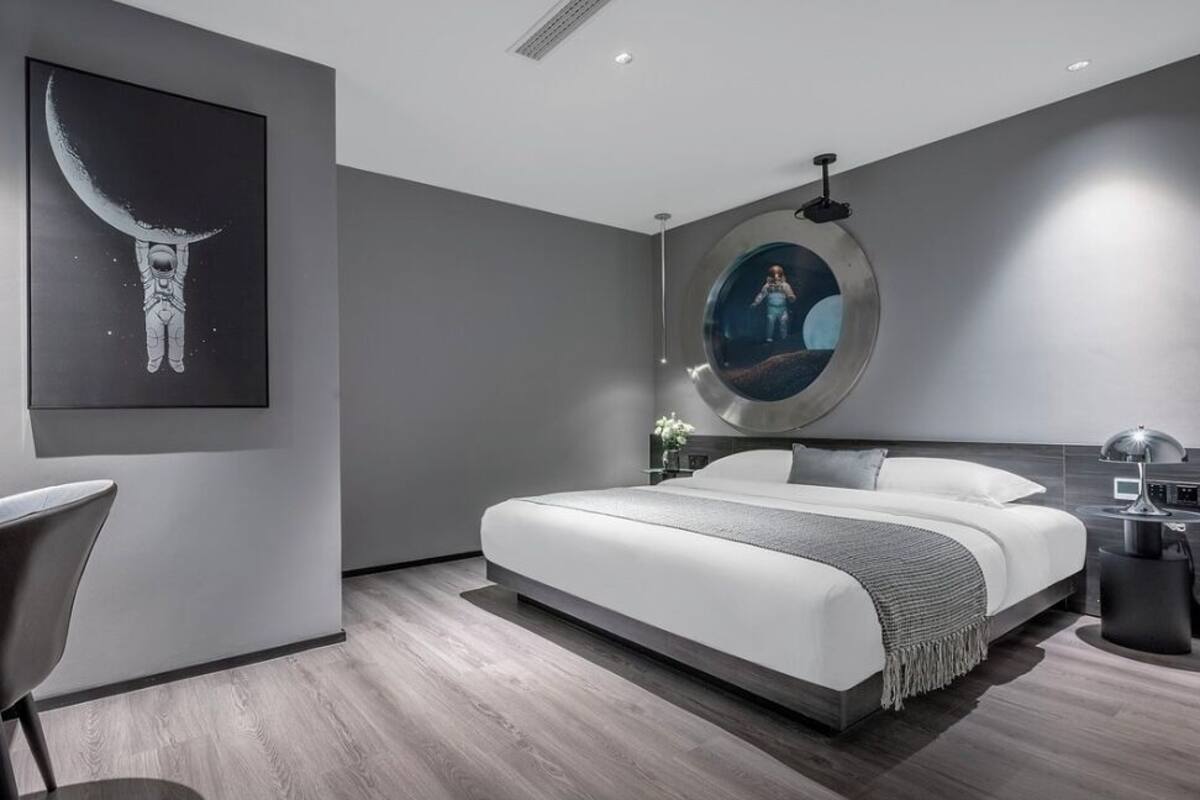 A modern bedroom with a large bed, a desk, and a chair. There is a painting of an astronaut on the wall and a round mirror above the bed.
