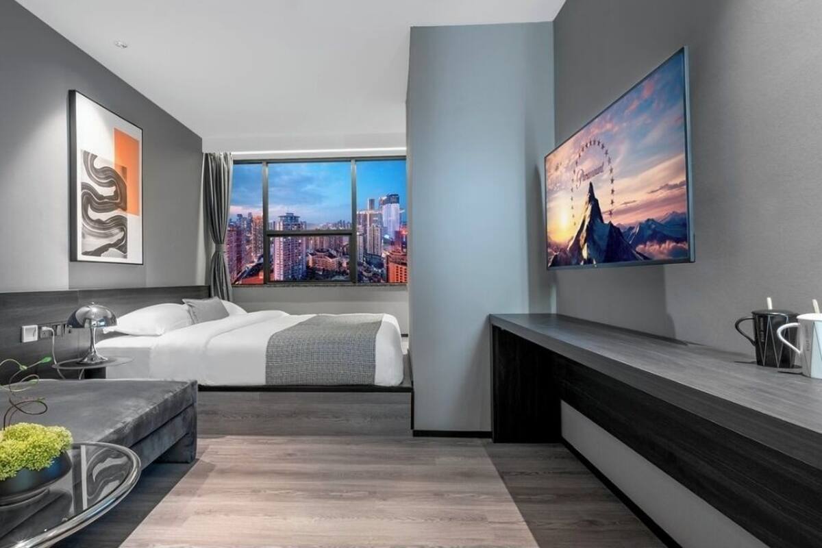 A modern hotel room with a bed, a desk, a large window with a city view, and artwork on the walls.