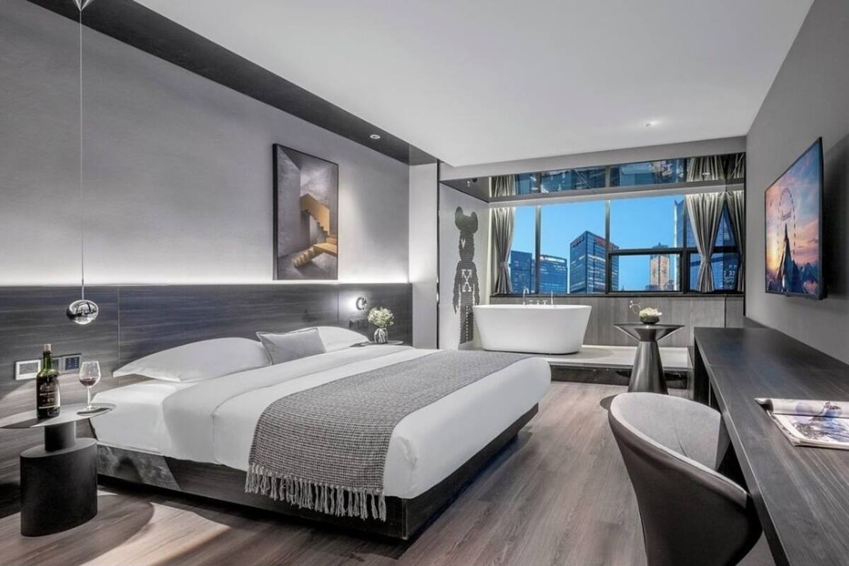A modern hotel room with a large bed, a sitting area with a chair, a desk, a bathtub, and a large window offering a city view.