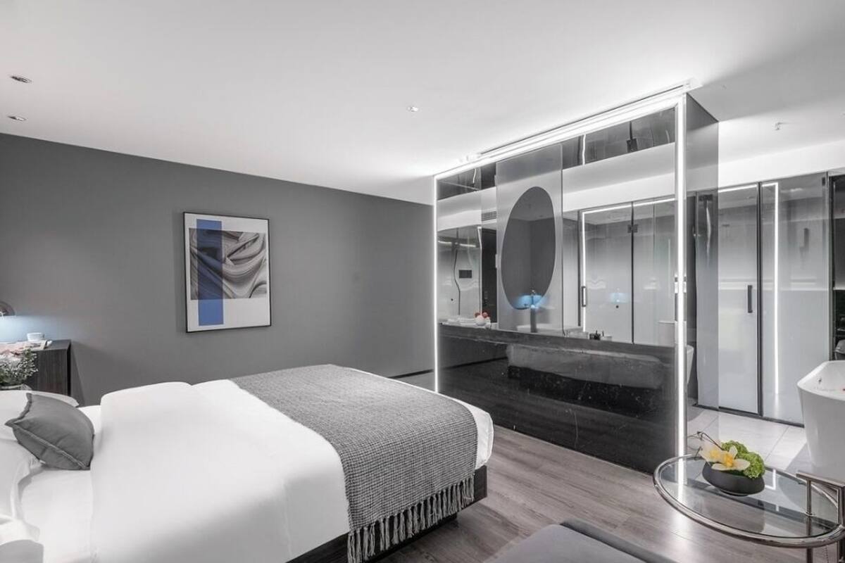 A modern hotel room with a large bed, a glass-enclosed walk-in closet, a separate bathroom with a freestanding bathtub, and a small round table with a flower vase.