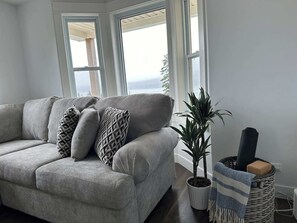 A living room with a grey sofa, patterned cushions, a potted plant, and a woven basket with a rolled-up mat.