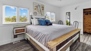 A bedroom with a bed, a nightstand, a wooden bench, a wall-mounted lamp, and a window with blinds.