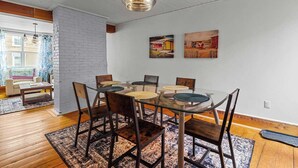 A dining area with a glass table and chairs, a hanging light fixture, and two paintings on the wall.