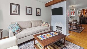 A living room with a sofa, coffee table, and fireplace. There are framed pictures on the wall and a TV mounted above the fireplace.