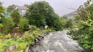 Un fiume che scorre attraverso un paesaggio verde e rigoglioso, con una casa sulla sinistra e una costruzione con tetto rosso sullo sfondo.