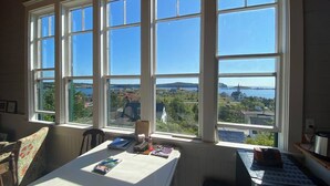 A room with a large window overlooking a scenic landscape, a table with various items, and a chair. - Trinity's Oceanview Gem: 2BR St. Paul's Schoolhouse (Trinity)