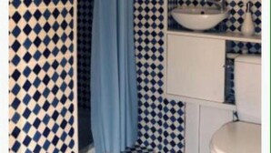 Un baño con suelo de baldosas a cuadros, cortina de ducha azul, lavabo con espejo y retrete blanco.