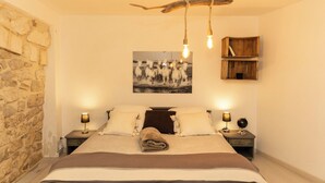 A bedroom with a stone wall, a bed with a brown blanket, two bedside tables with lamps, and a rustic wooden shelf. - 🌿 Instant en Provence - LE GARDIAN Air-conditioned apartment in the heart of Alpilles (Eyragues)