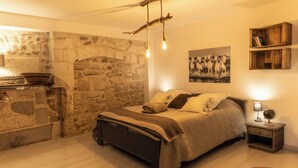 A cozy bedroom with a stone wall, a bed with a headboard, a nightstand with a lamp, and a shelf with books. - 🌿 Instant en Provence - LE GARDIAN Air-conditioned apartment in the heart of Alpilles (Eyragues)