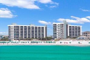 A beachfront hotel with multiple floors, balconies, and a sandy beach area with umbrellas.