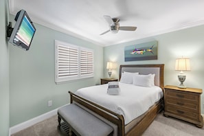 A bedroom with a bed, a bench, a ceiling fan, two bedside tables with lamps, a TV mounted on the wall, and a window with blinds. - Mainsail 471 - Gulf Front Condo, Community Pool, Hot Tub, Free Beach Service! (Miramar Beach)