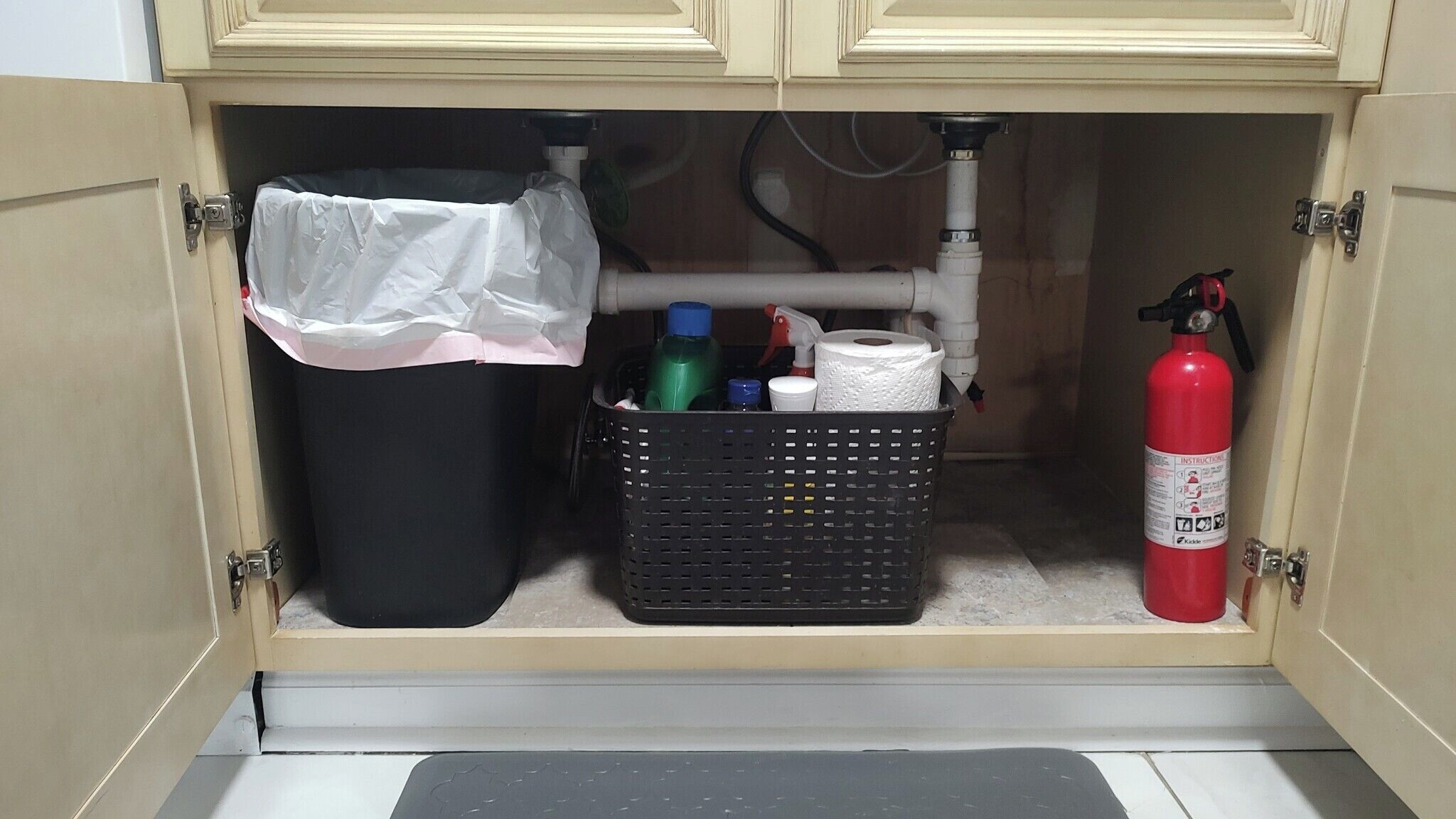A utility cabinet with a black trash bin, a black basket containing cleaning supplies, and a red fire extinguisher.
