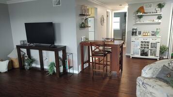 Een woonkamer met een flatscreen televisie, een houten eettafel en stoelen. Er is een witte kast met glazen deuren, waarin keukengerei staat.