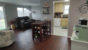 Una sala de estar con un sofá de cuero oscuro, una mesa de comedor de madera y una cocina con armarios de color claro y un electrodoméstico verde.