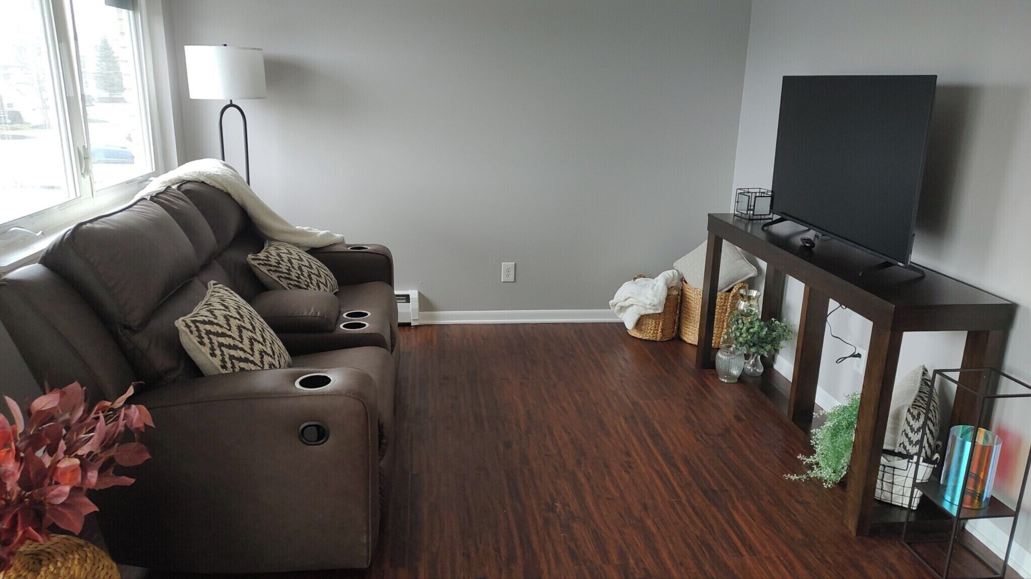 A modern living room with a brown leather sofa, a flat-screen TV on a wooden stand, and a large window providing natural light.
