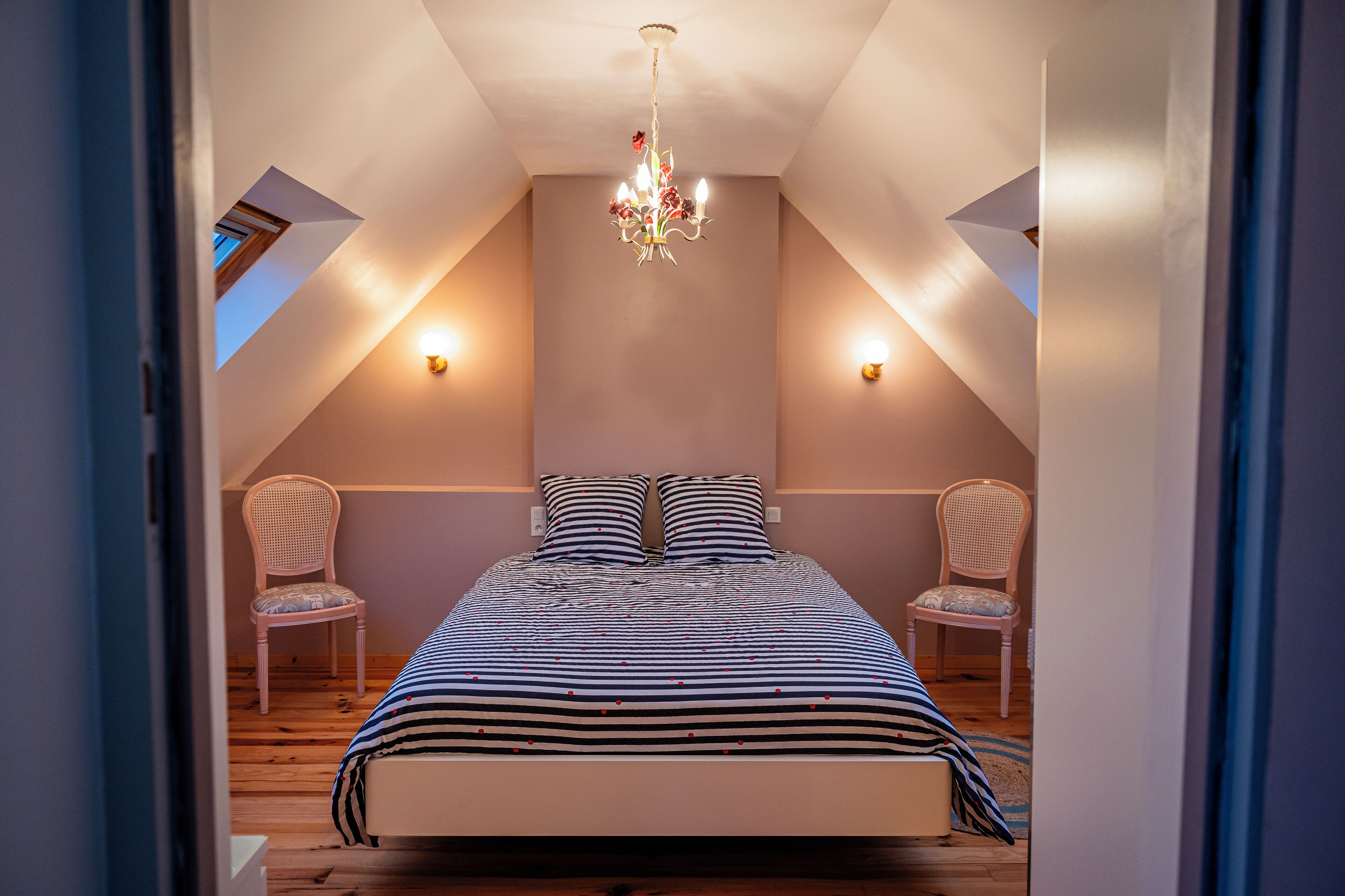 Une chambre avec un plafond en pente, un lit avec une literie à rayures, deux chaises et un lustre.