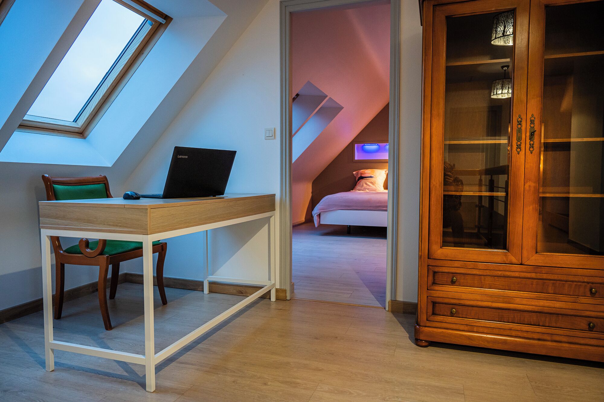 A room with a sloped ceiling, a desk with a laptop, a chair, a bed, and a wooden cabinet.