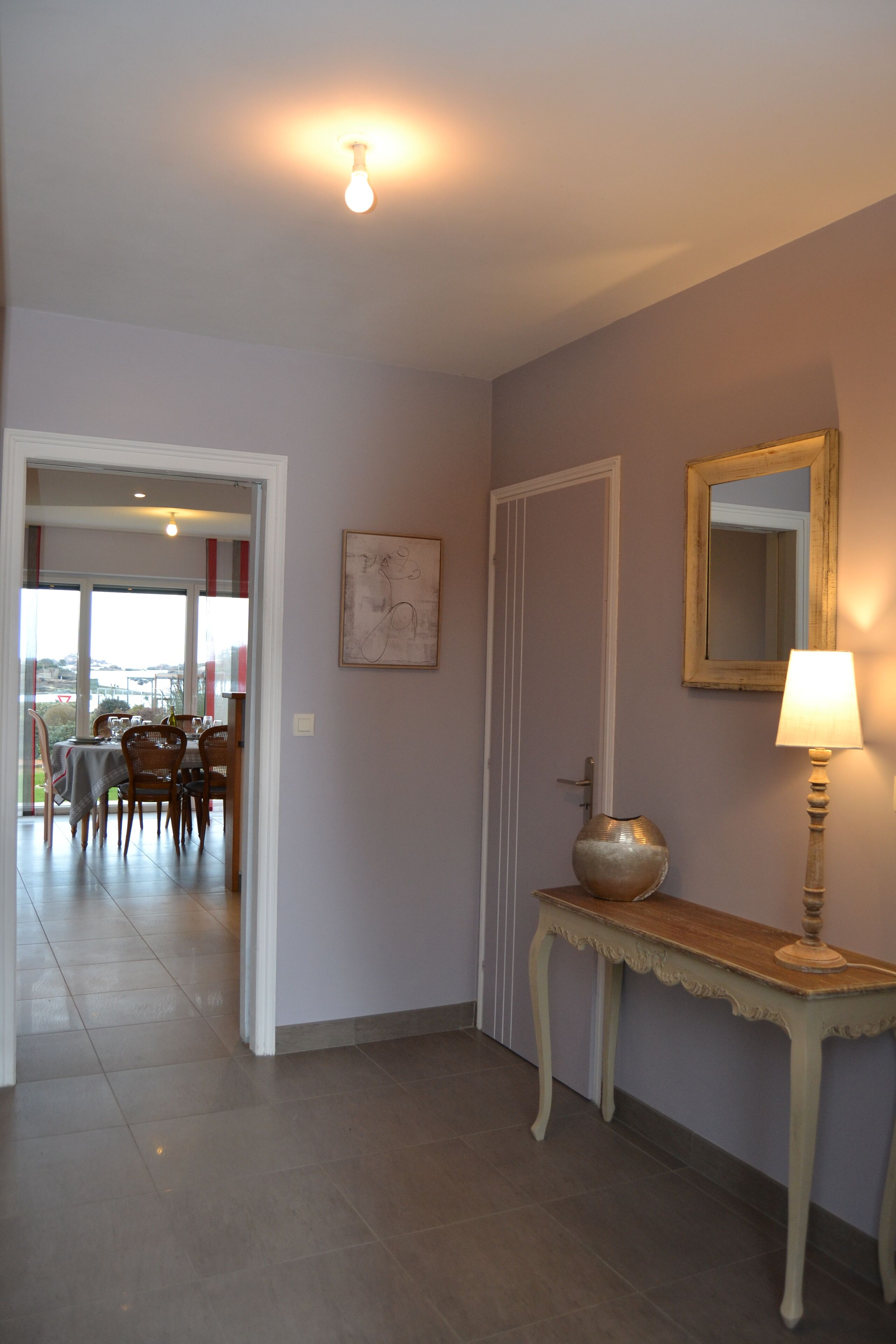 A hallway with a dining area in the background, a door, a mirror, and a table with a lamp.