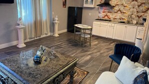 Una sala de estar con una mesa de centro de mármol, un sofá azul y una cocina con armarios blancos y un refrigerador negro.