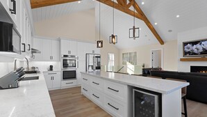 A modern kitchen with white cabinetry, a large island, and a built-in oven. There is a dining area with a black sofa and a flat-screen TV mounted on the wall.
