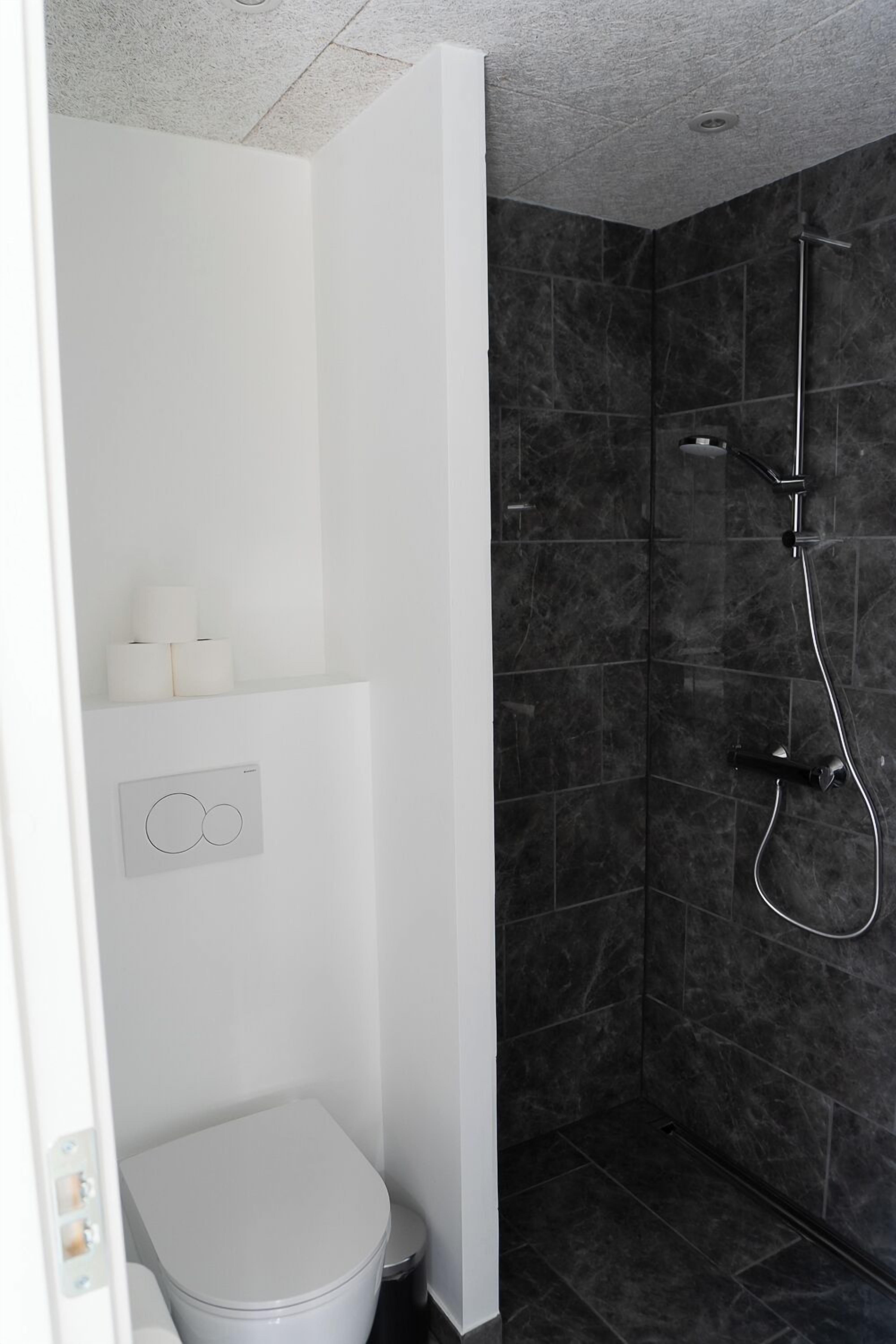 A modern bathroom with a white toilet, a shower with dark tiles, and a wall-mounted light switch.