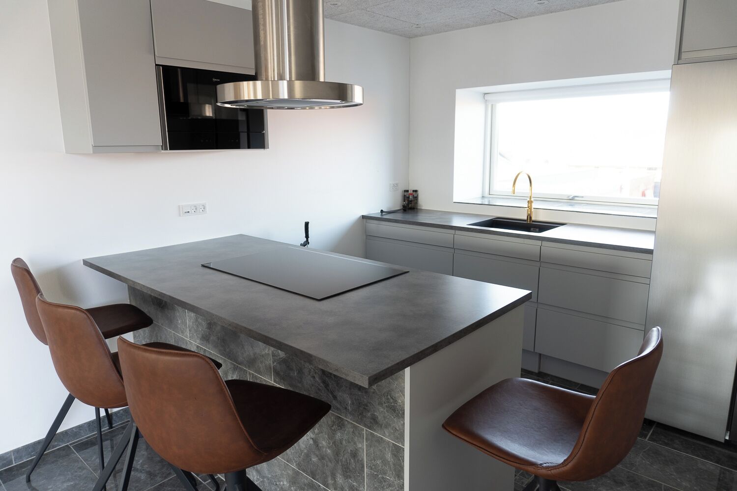 Una cocina moderna con una isla central, dos sillas de cuero marrón, una campana extractora de acero inoxidable, una ventana con vistas y un horno empotrado.