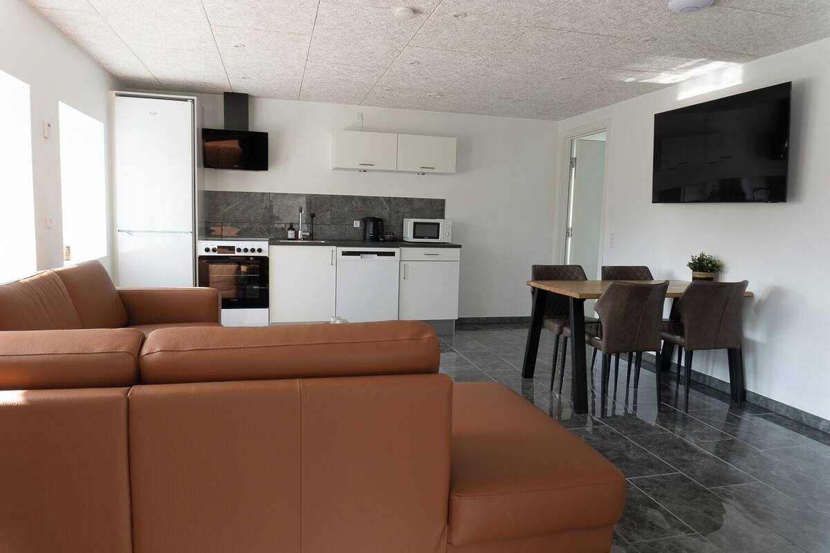 A modern living room with a brown sectional sofa, a dining area with a wooden table and chairs, a kitchen with white cabinets and appliances, and a flat-screen TV mounted on the wall.