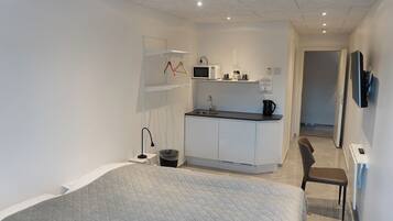 A compact bedroom with a bed, a small desk, a chair, a kitchenette with a microwave, and a TV mounted on the wall.