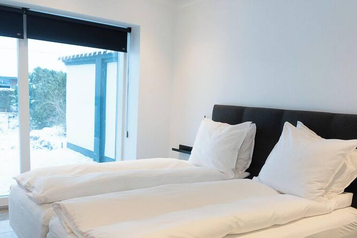 A double bed with white linens in a modern room with a large window and a sliding door.