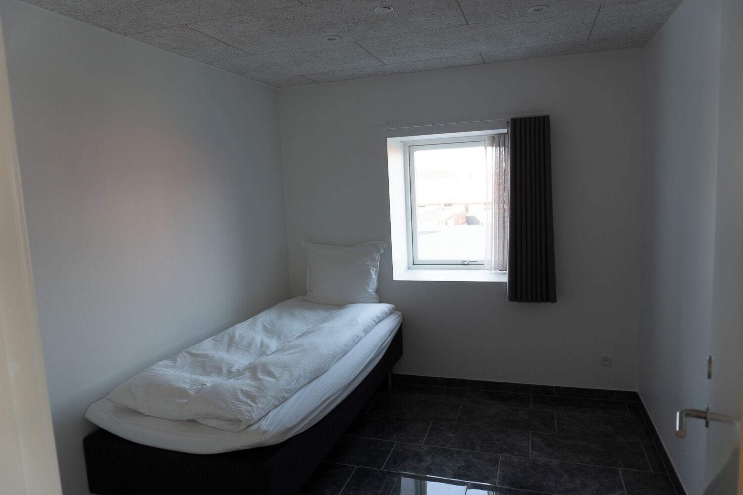 A small, narrow room with a single bed, a window with curtains, and a tiled floor.