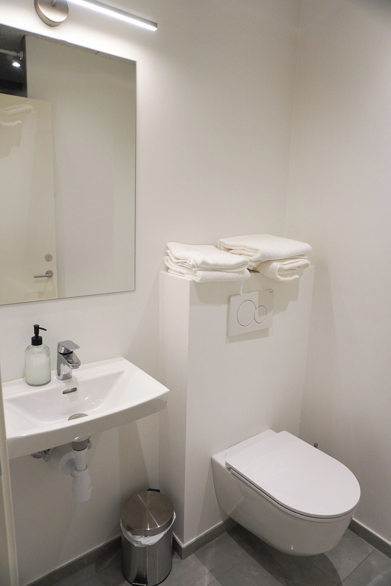 A modern bathroom with a white sink, mirror, and toilet. There are folded towels on a shelf above the toilet.