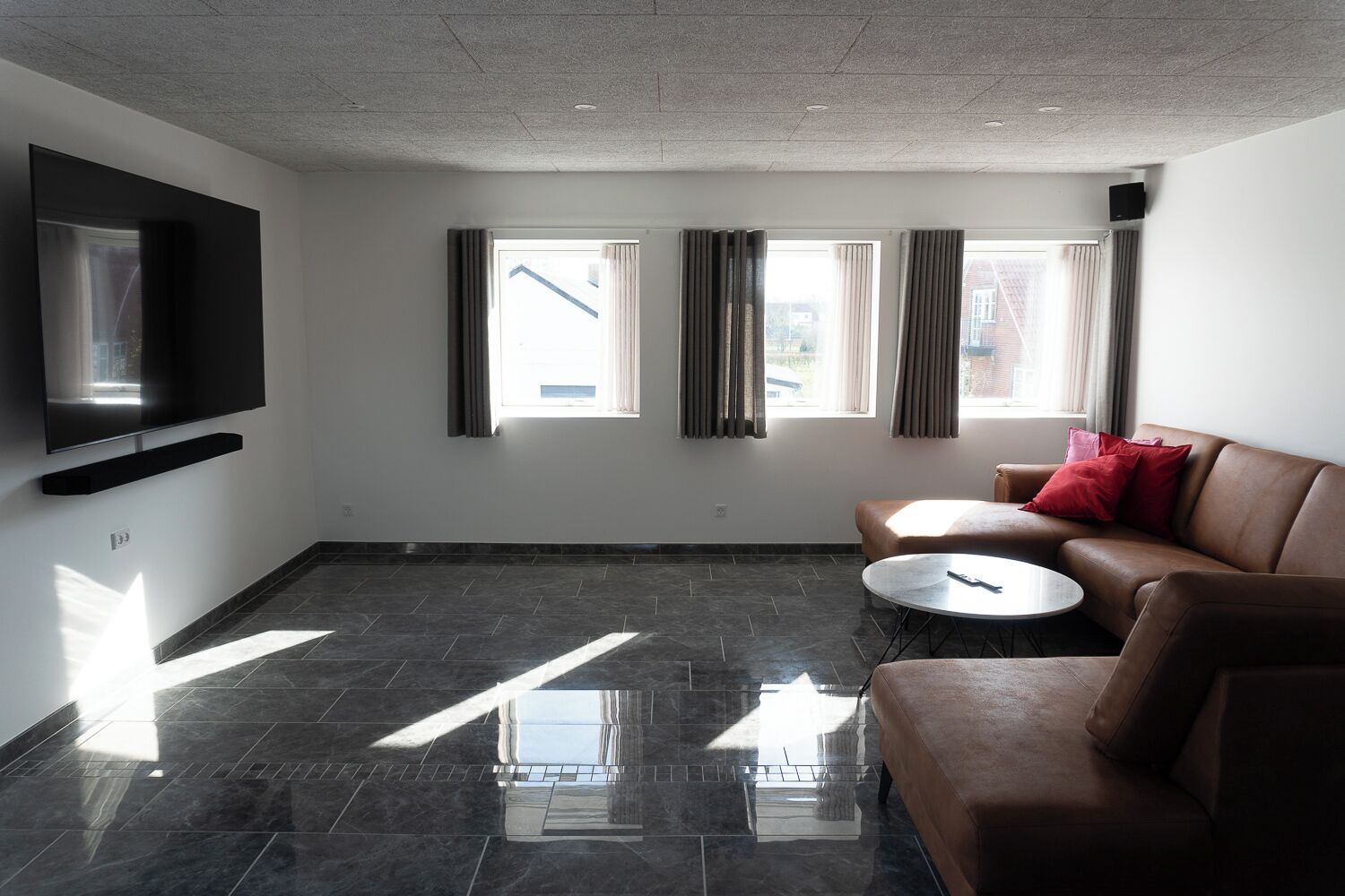Een moderne woonkamer met een flatscreen televisie aan de muur, een bruine leren bank, een ronde salontafel en een groot raam met gordijnen.