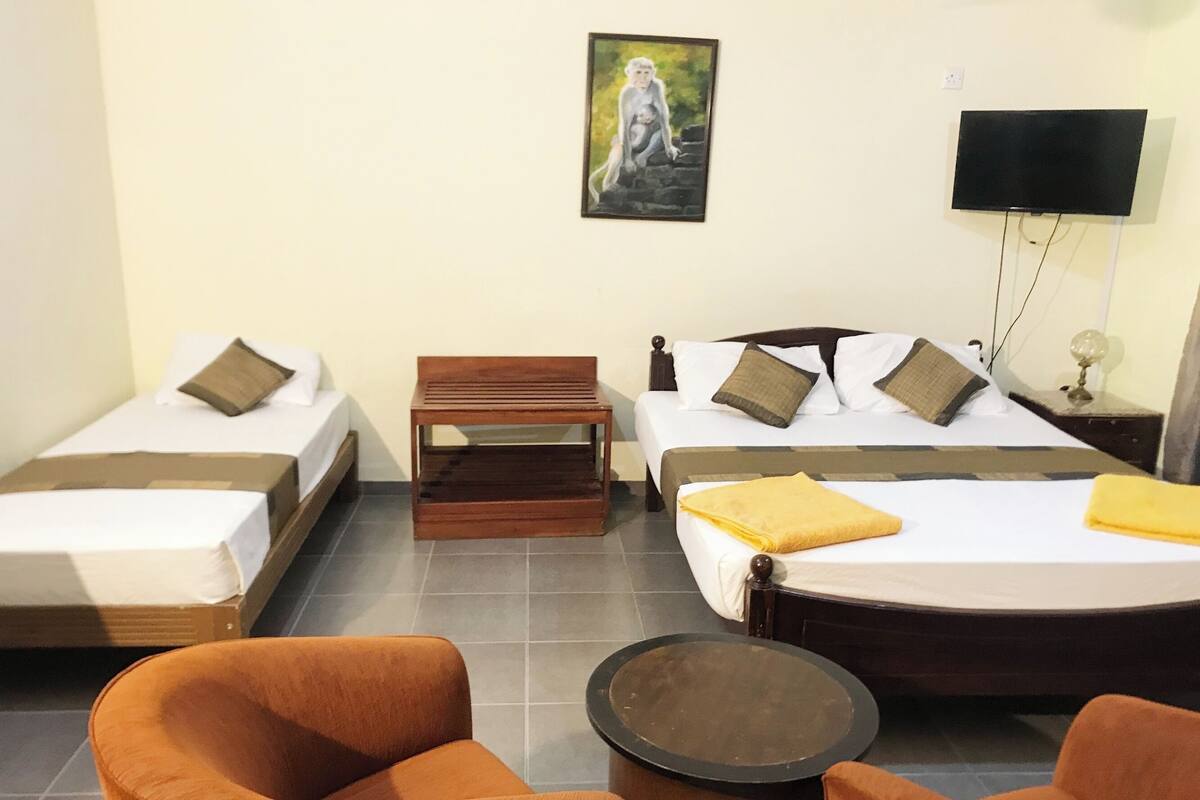 A hotel room with two single beds, a double bed, a wooden bench, two orange armchairs, and a small round table.