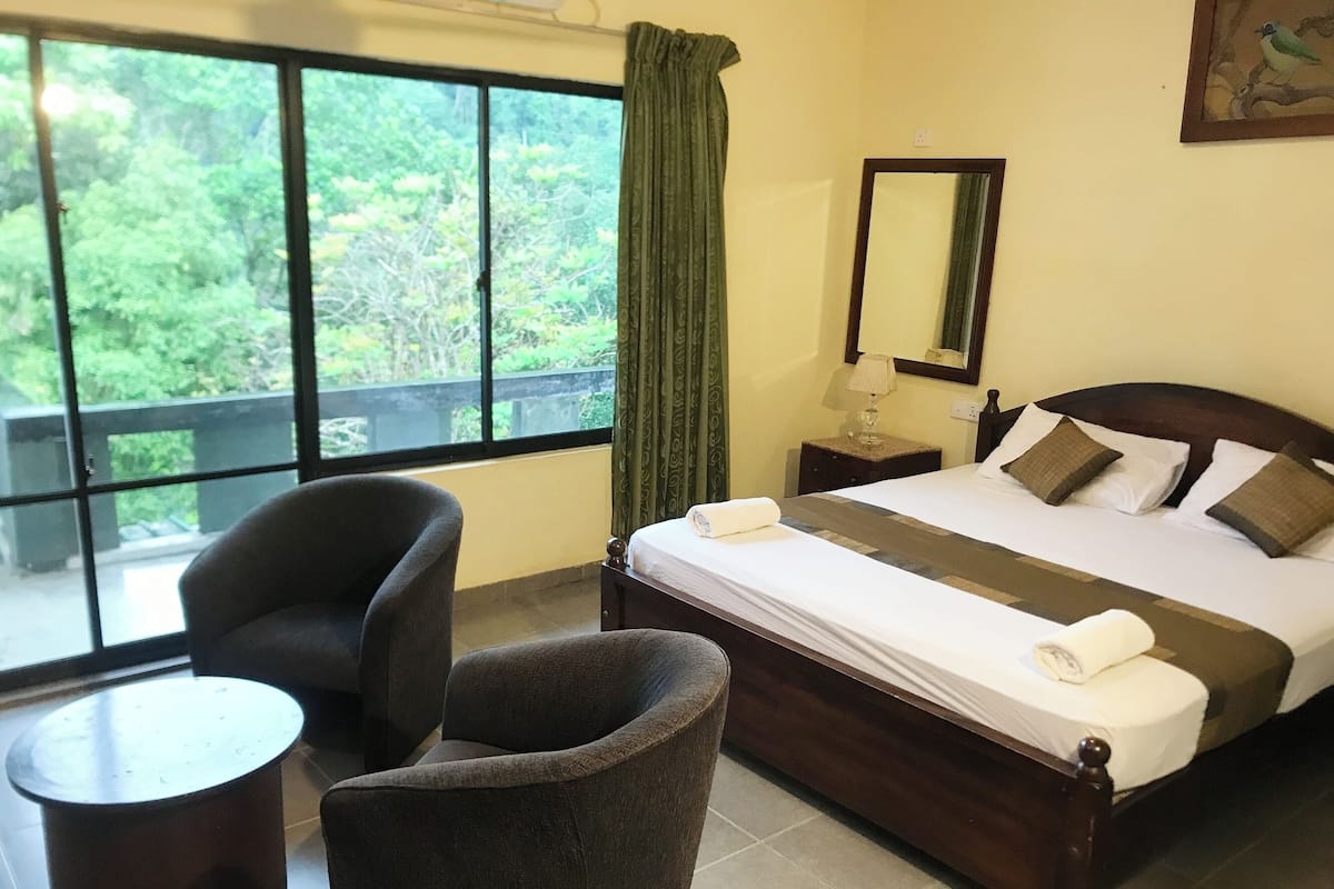A hotel room with a large window, a bed with pillows, a chair, a small table, and a nightstand with a lamp.