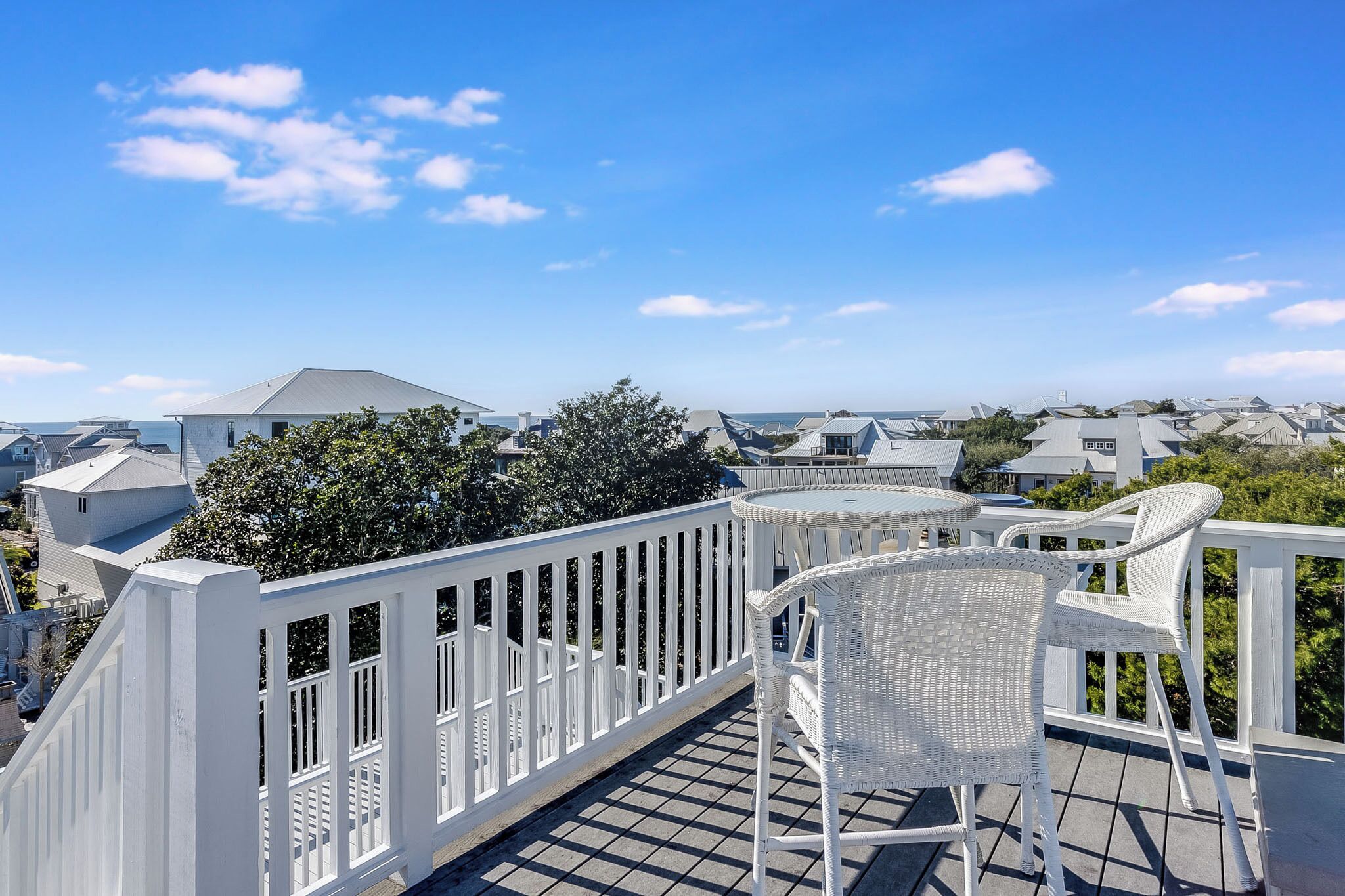 A balcony with white wicker furniture and a white railing, overlooking a residential area with houses and trees.