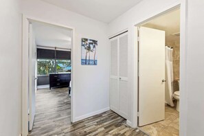 A hallway with a wooden floor, a door leading to a bathroom, and a view of a bedroom with a bed and a window. - Partial Oceanview 2 bedroom condo, across st from ocean, walk to shops & dining (Napili-Honokowai)