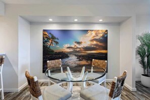 A dining area with a glass table surrounded by chairs, a large painting of a sunset over a beach, and a potted plant. - Partial Oceanview 2 bedroom condo, across st from ocean, walk to shops & dining (Napili-Honokowai)